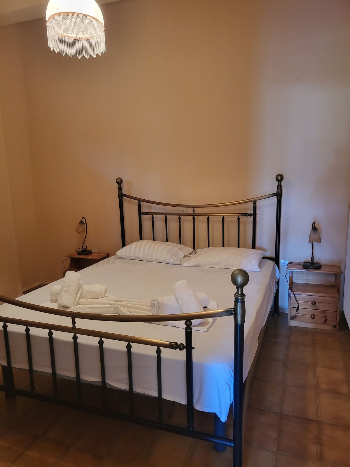 A cozy bedroom features a simple metal bed frame with a neatly made bed, complemented by fresh linens. A bedside table holds a lamp, while soft lighting enhances the relaxing atmosphere. The walls are painted in a neutral tone, contributing to a calm environment.