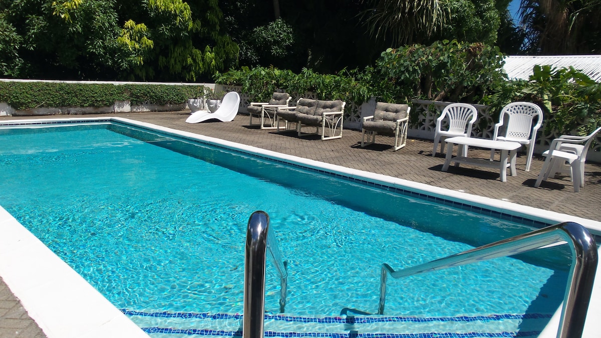 A refreshing swimming pool is surrounded by a paved deck, featuring several lounge chairs and sunbeds. Lush greenery and vibrant trees provide a natural backdrop, enhancing the sense of tranquility and relaxation in this open-air recreation area.