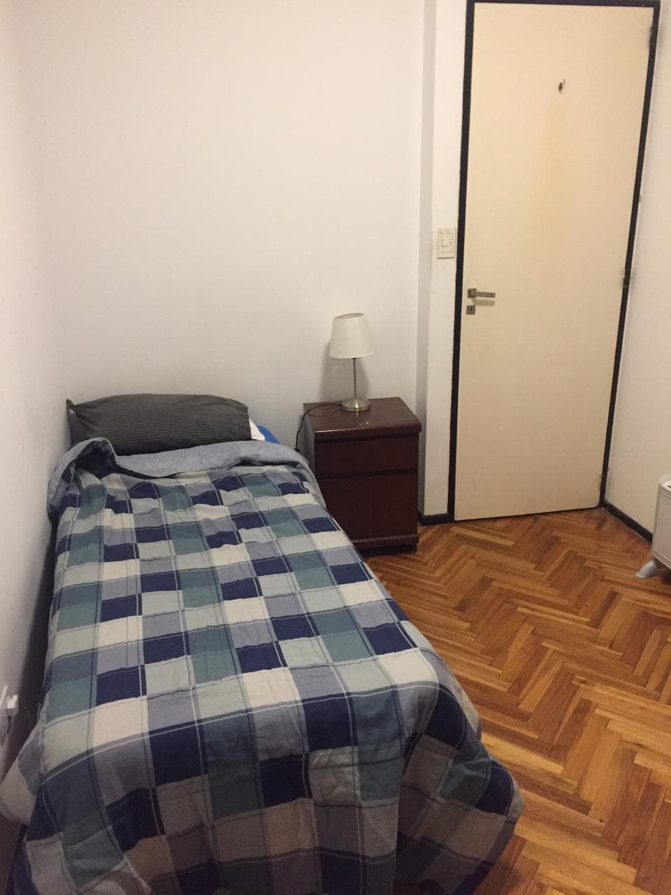 A small room features a narrow bed topped with a checkered blanket, accompanied by a bedside table with a lamp. The room is characterized by hardwood flooring and a plain white wall, with a door leading to the hallway.