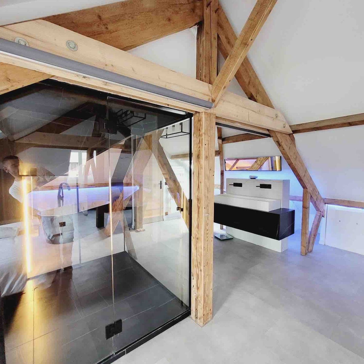 An open space design features exposed wooden beams against a bright ceiling. A modern glass shower enclosure is visible, alongside a sleek white and black vanity unit, providing a minimalist aesthetic. Natural light enhances the spacious feel, flowing into the well-lit area.