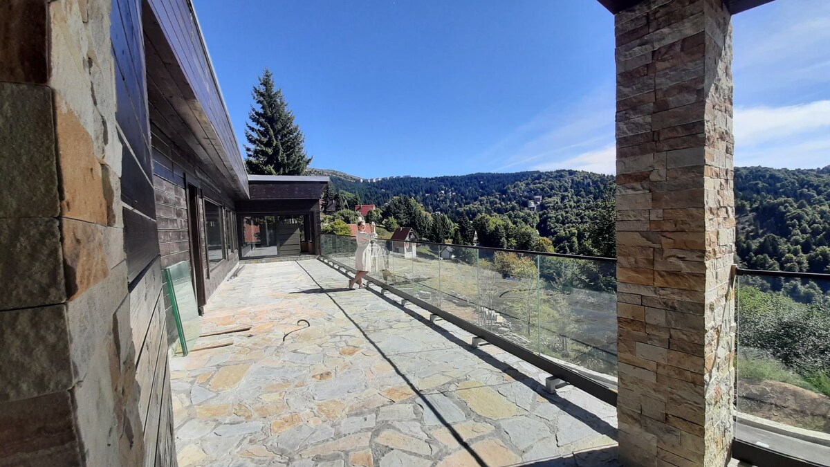 A spacious outdoor terrace is presented, featuring natural stone flooring and glass railings. The surrounding landscape offers a view of lush greenery and distant hills under a clear blue sky, creating a calm atmosphere for relaxation.