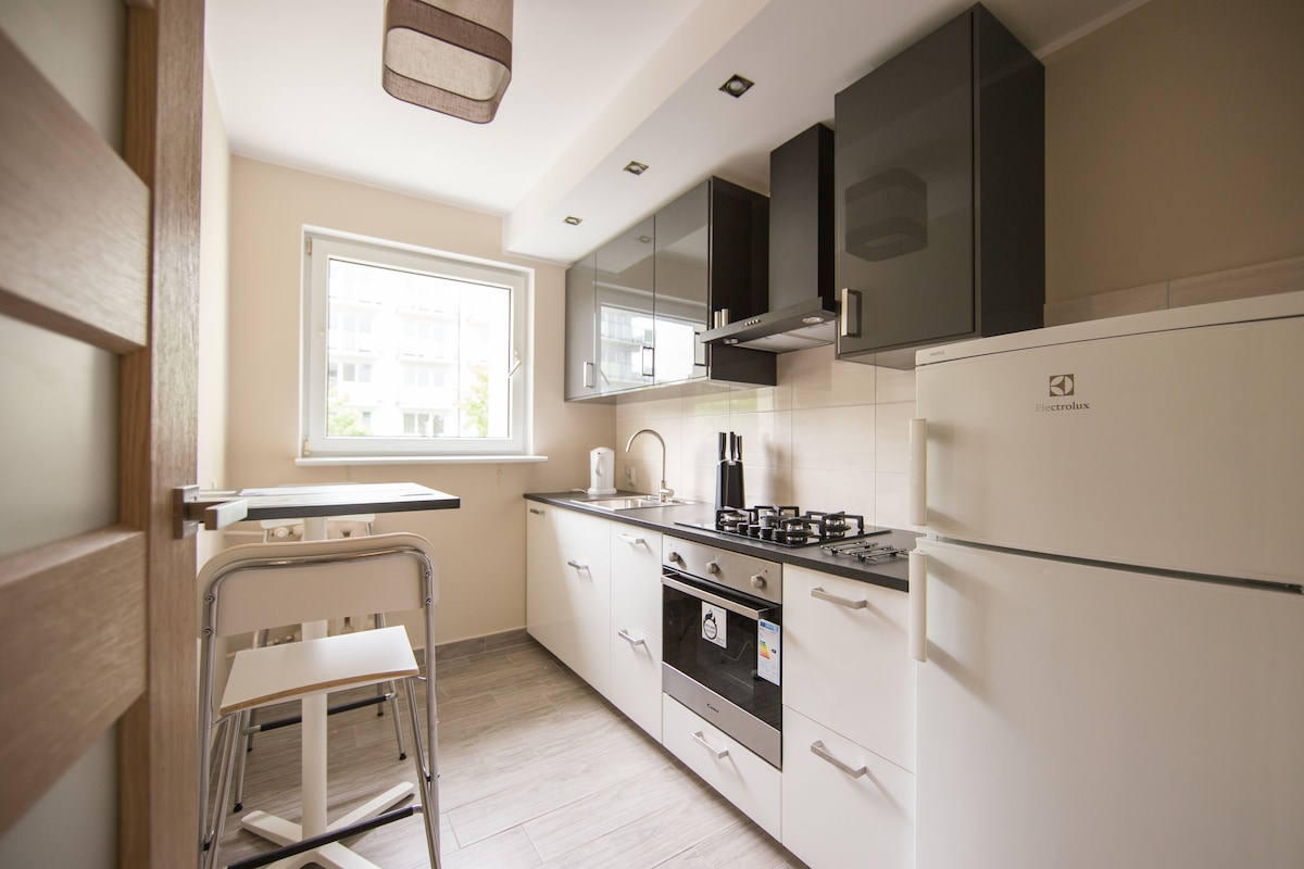 A modern kitchen features sleek cabinetry with a mix of dark and light finishes. A window allows natural light, highlighting a compact dining table with two high stools. Appliances, including a gas stove and refrigerator, enhance functionality.