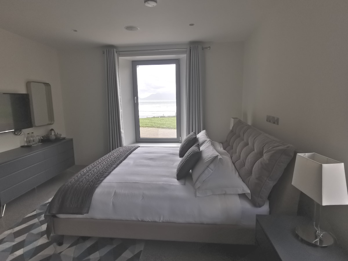 A spacious bedroom features a large bed with plush gray accents and neatly arranged pillows. A modern dresser is situated across from the bed, while a window with sheer curtains offers a view of the beach outside, filling the room with natural light.