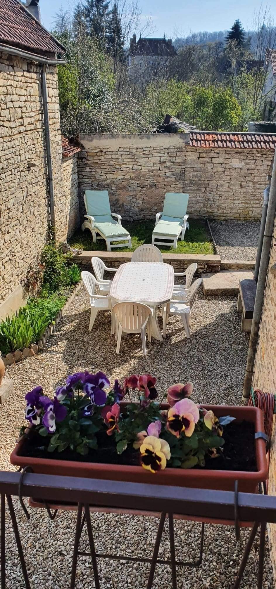 A gravel courtyard area is presented, featuring a round dining table surrounded by chairs. Two lounge chairs are positioned for relaxation. Potted flowers add color, while a small grassy section is included. Stone walls and greenery create a serene outdoor space.