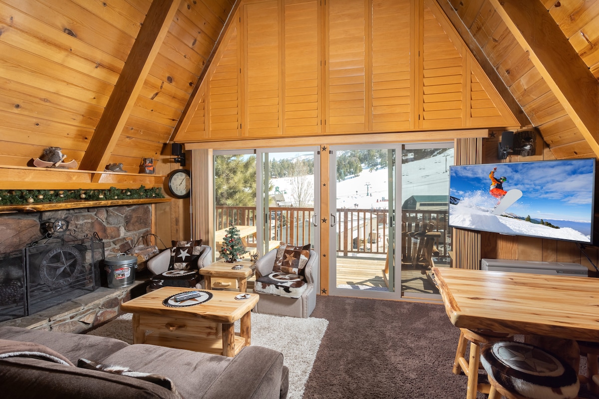 A welcoming living area showcases a high ceiling with exposed wooden beams. A comfortable seating arrangement includes a plush sofa and wooden coffee tables. Large glass doors provide access to a deck, while a flat-screen TV displays a winter sports scene, enhancing the chalet's cozy ambiance.