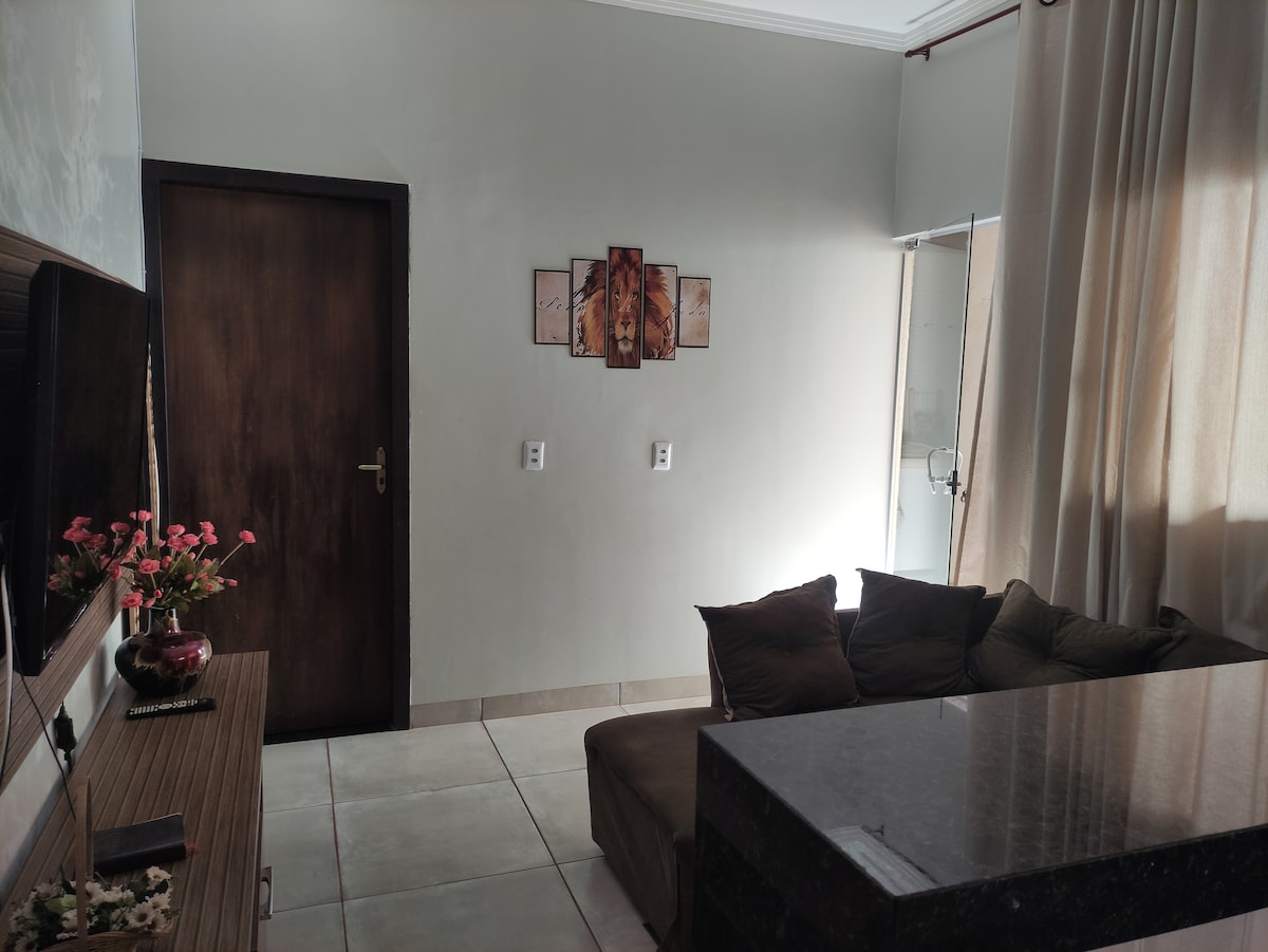 A living area is shown featuring a dark brown door and a large window covered with light curtains. A dark sofa sits against the wall, complemented by a coffee table made of black material. Wall art depicting three abstract panels is displayed above the sofa.
