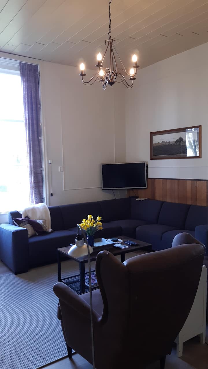 Uw woonkamer is groot met een hoog plafond, kroonluchter, televisie, zitbank en 2 grote leesstoelen die uitzicht bieden op de grote tuin en weiden. 