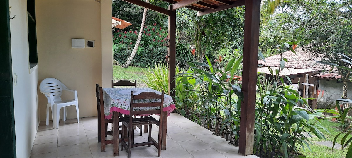 A covered outdoor patio features a wooden table set with chairs, surrounded by lush greenery. The tiled floor enhances the space, with a glimpse of a well-maintained garden visible in the background.