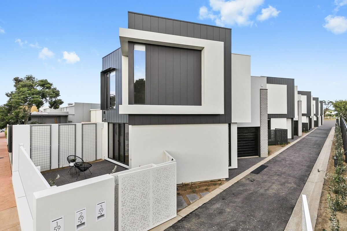 The modern townhouse features a sleek façade with a combination of dark and light materials. A small outdoor seating area is visible in the foreground, providing a welcoming entrance. The surrounding area is clear, highlighting the clean lines and architectural design.
