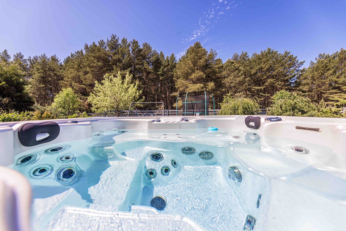 A hot tub is situated outside, surrounded by lush green vegetation and tall pine trees, with clear water reflecting the sky. The relaxing space offers multiple jets, creating a tranquil atmosphere in a natural setting.