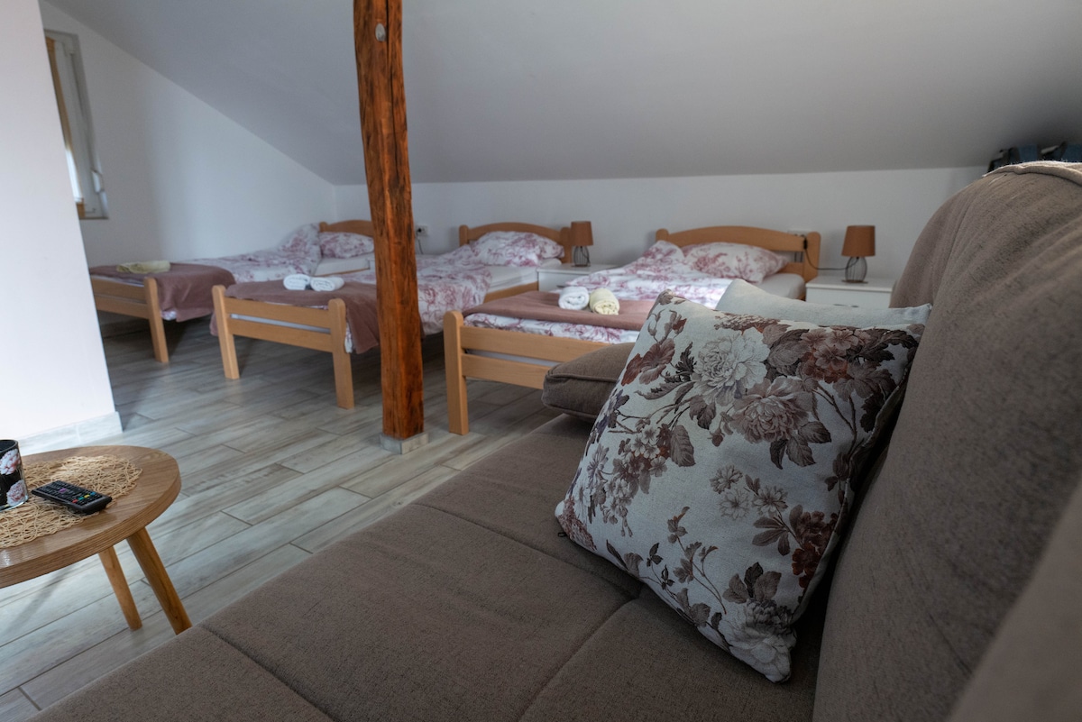 A spacious studio room is shown with multiple beds arranged in a cozy layout. Wooden beams add character to the sloped ceiling. A comfortable sofa and decorative pillow offer seating, while bedside lamps are positioned next to each bed.