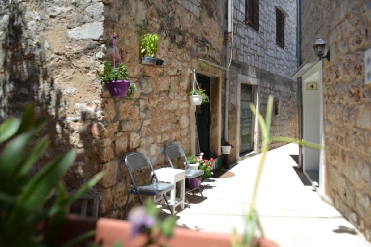 A narrow stone street is captured, lined with rustic buildings featuring textured walls. Small potted plants decorate the entrance of a doorway. Two metal chairs and a side table are positioned against the wall, inviting a quiet moment outdoors.