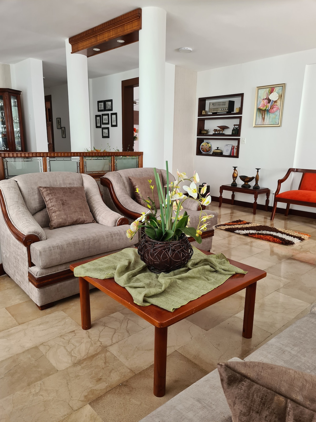 A bright living area features two plush armchairs and a stylish sofa, arranged around a wooden coffee table. A decorative plant sits at the center of the table, and art pieces are displayed on the walls, enhancing the welcoming ambiance of the space.