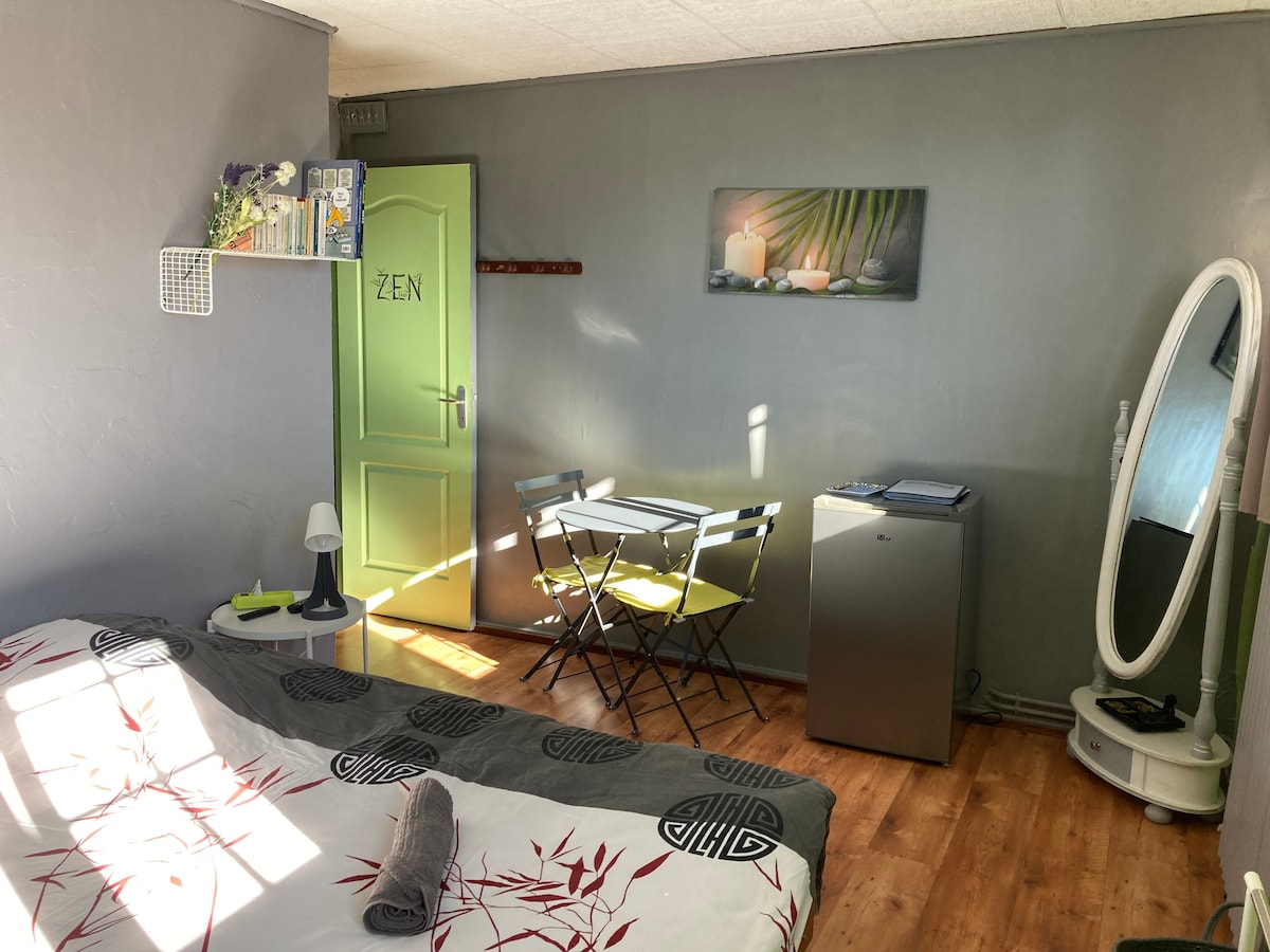 A spacious room features a large bed with decorative bedding, a round table with two folding chairs, and a mirrored cabinet. A small fridge is positioned in one corner, while natural light enters through a nearby window, highlighting the clean lines and calm colors of the decor.