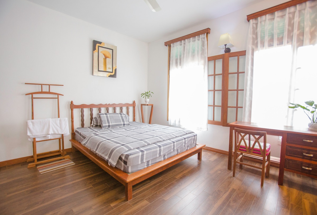 A spacious bedroom is furnished with a wooden bed featuring a plaid-patterned blanket. Natural light filters through two large windows, accented by sheer curtains. A wooden desk and chair are positioned beside a small indoor plant, with a traditional wooden drying rack nearby.