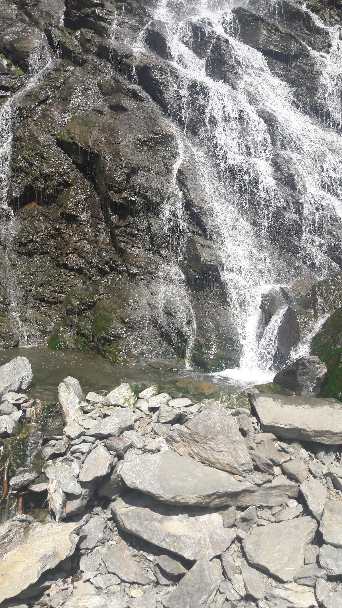 A cascading waterfall flows over rugged rocks, creating a serene atmosphere. The clear water tumbles down the rocky surface, with a small pool forming at the base. Various textured stones are scattered in the foreground, reflecting the natural beauty of the surrounding area.