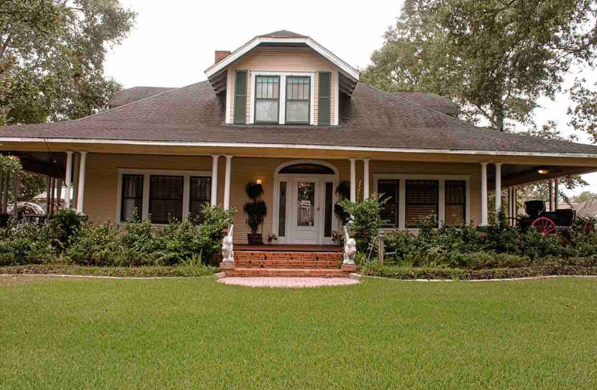 A charming exterior of a two-story house is presented, featuring a wraparound porch supported by white columns. Lush green grass surrounds the property, while well-maintained bushes and flower beds enhance the inviting entrance. The roof has a distinctive gable design, adding to its character.