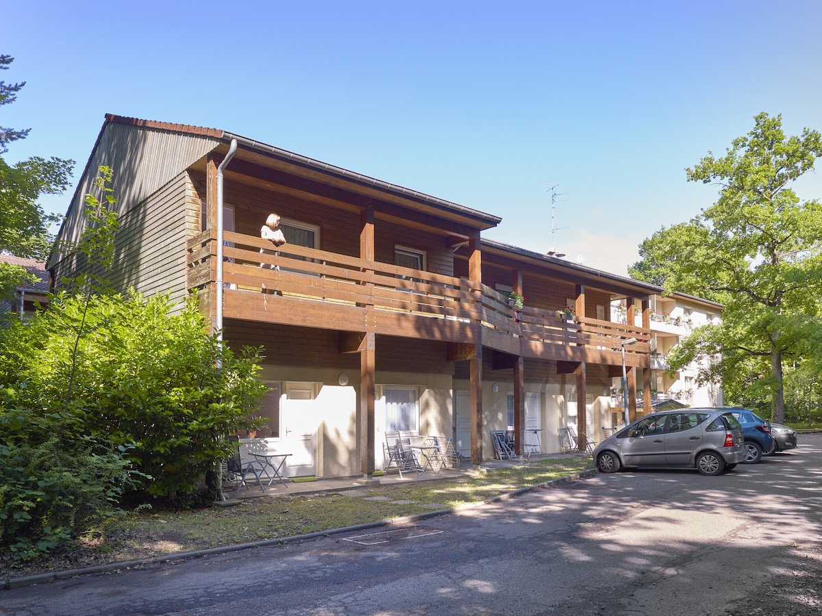 The two-story chalet features wooden balconies with seating arrangements, surrounded by greenery. The entrance doors are visible, leading to the individual units. A small lot is situated in front, accommodating parked vehicles, while trees provide a natural backdrop.