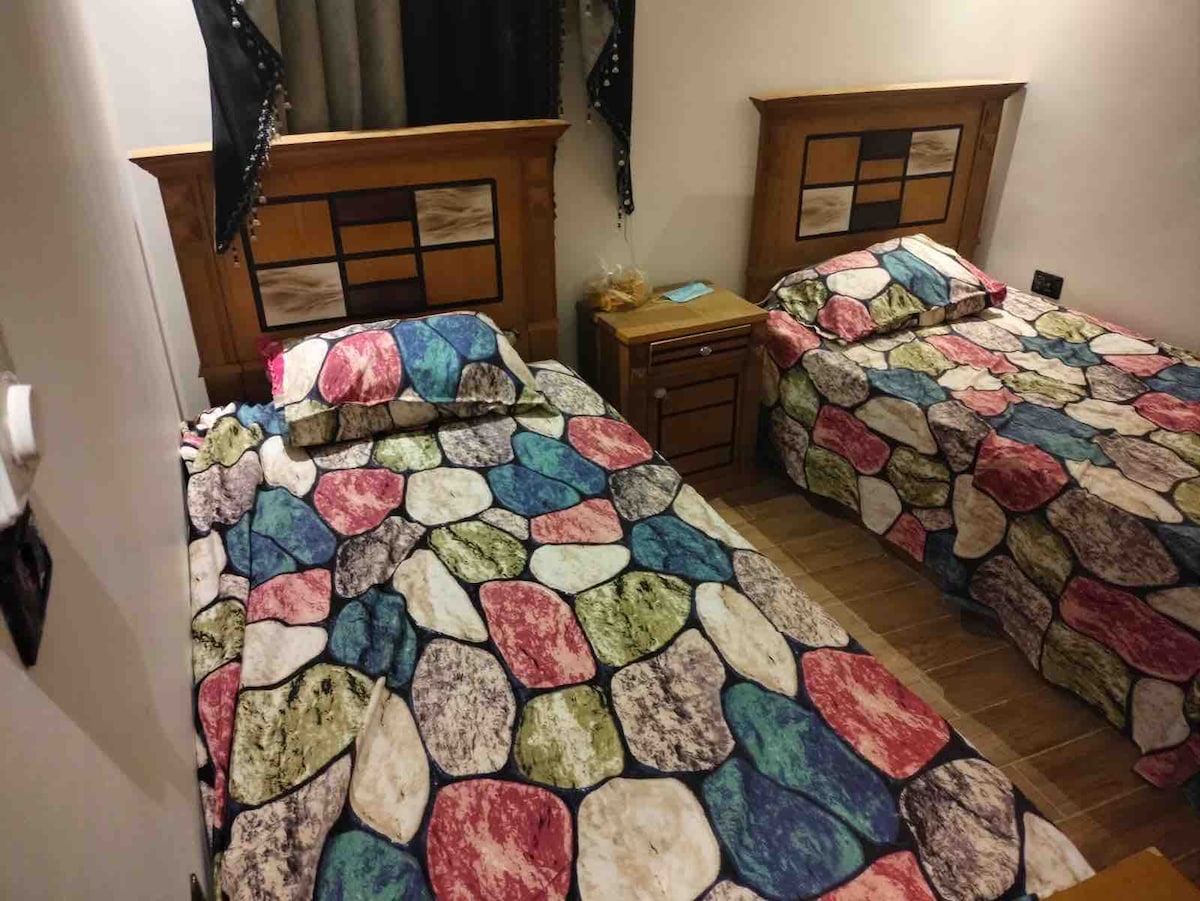 The room features two spacious beds, each adorned with colorful rock-patterned bedding. A bedside table is positioned between the beds, providing convenience. Soft lighting illuminates the space, while curtains frame the windows, creating a cozy environment.