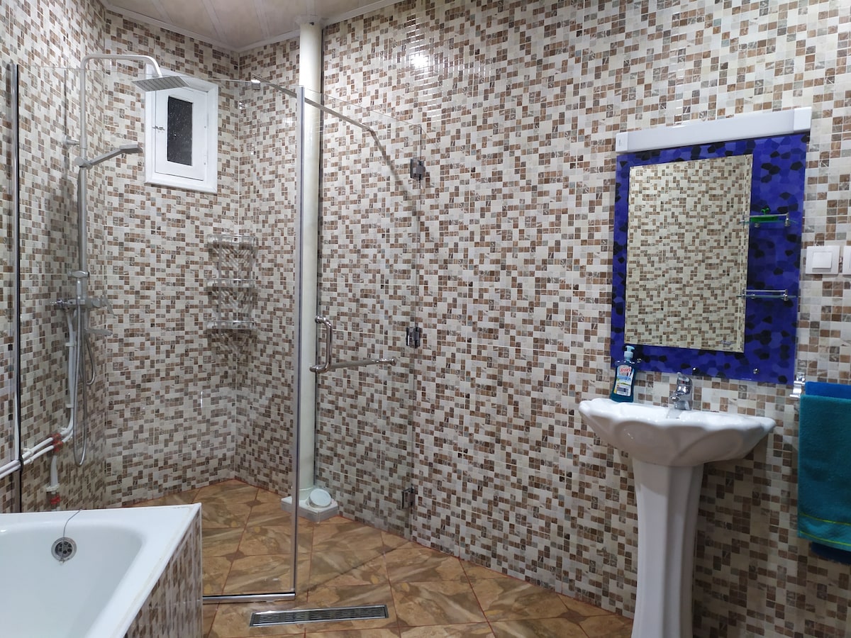 A modern bathroom features a walk-in shower with a glass enclosure and a bathtub. Textured tiles cover the walls and floor, while a sink is positioned beside a large mirror bordered by a blue frame. A towel hangs nearby for convenience.
