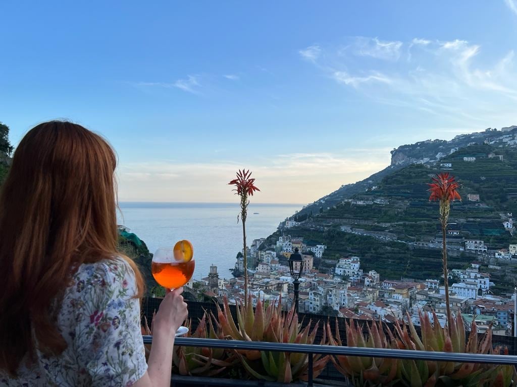 A scenic view of the coastline and hillside village is visible from a terrace. Bright orange flowers frame the scene, while a person is seen holding a drink and gazing towards the horizon, where the sea gently meets the sky.