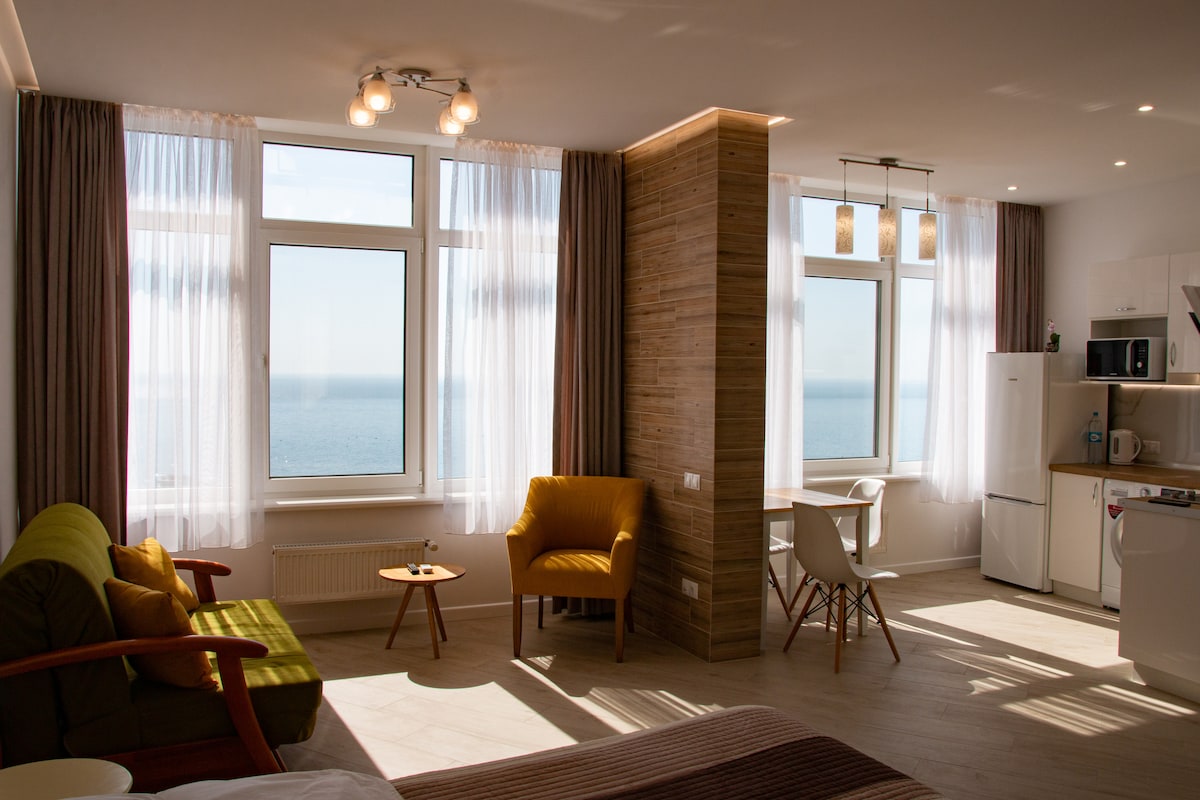 Bright and airy living space features large windows providing an unobstructed view of the sea. A cozy sofa and armchair are arranged around a small coffee table. The kitchenette is equipped with modern appliances, and a dining area is visible, illuminated by natural light.