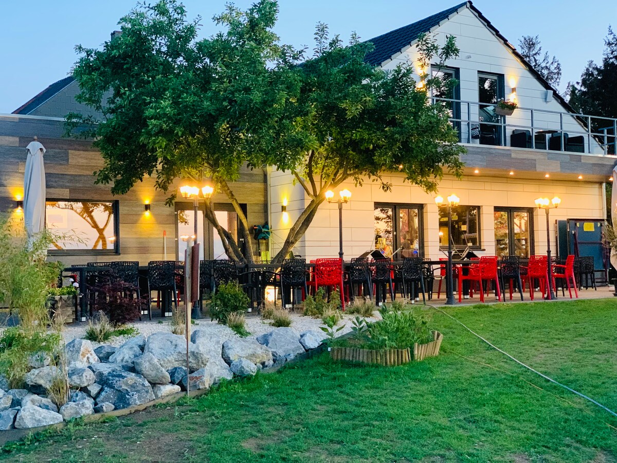 The exterior view of a modern building showcases a spacious patio area with multiple red and black chairs arranged around tables. Soft lighting illuminates the area, enhancing the ambiance. Lush greenery and stone features are present in the foreground, contributing to a pleasant outdoor setting.