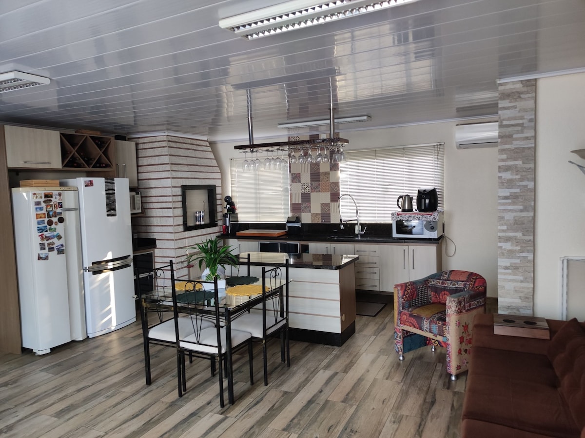 The spacious kitchen and living area features modern appliances, including a white refrigerator and a coffee maker. A dining table with black chairs is complemented by a colorful armchair. Large windows allow natural light to fill the space, highlighting the sleek countertops and patterned backsplash.