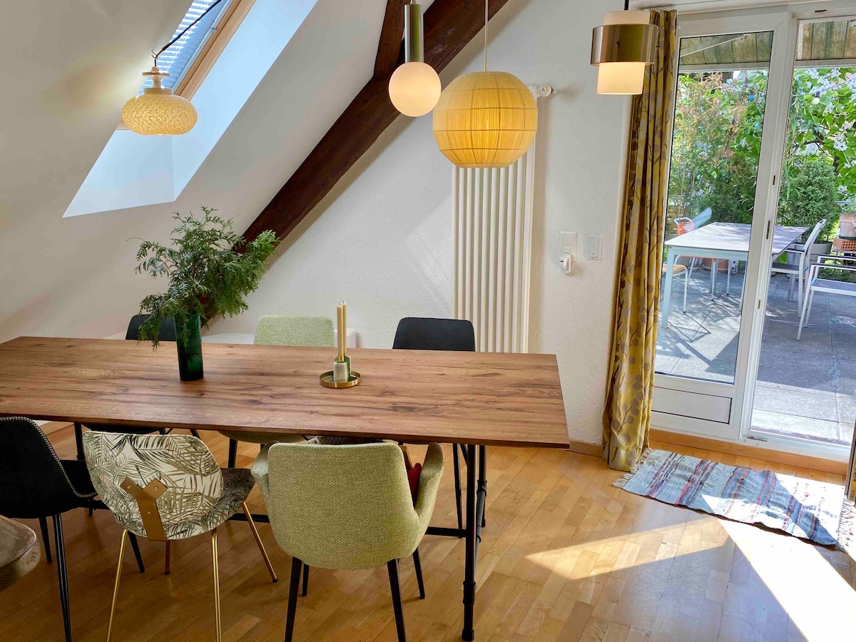 A dining area is highlighted by a large wooden table surrounded by colorful chairs. Natural light pours in through a slanted window, illuminating a green vase with greenery. A glass door leads to a spacious balcony, enhancing the open feel of the space.