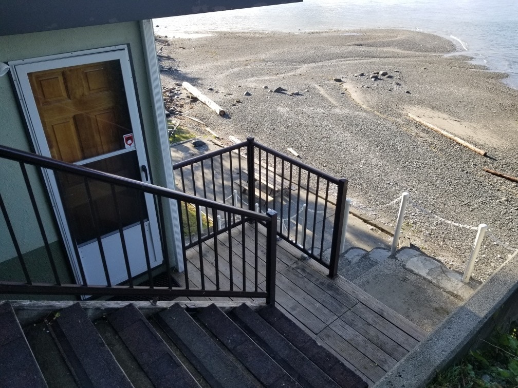 A set of wooden stairs leads down to a sandy beach area, with pebbles and driftwood visible along the shoreline. The entrance door is partially visible at the top of the stairs, framed by a railing that offers a clear pathway to the water's edge.