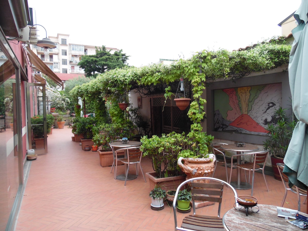 An inviting courtyard is filled with lush greenery and potted plants. Several round tables and chairs are arranged throughout the space, surrounded by vibrant murals. The area offers a peaceful retreat for guests to relax and socialize, with an open and airy atmosphere.