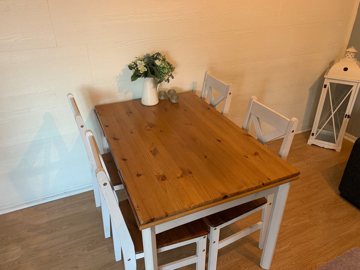 A wooden dining table is set against a softly lit wall. Four white chairs surround the table, one of which is positioned slightly apart. A small vase containing greenery decorates the center, adding a simple touch to the space.
