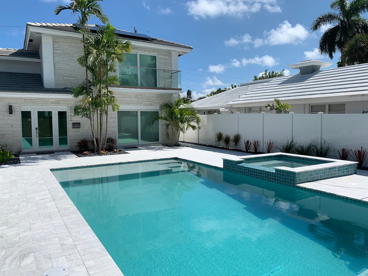 A bright outdoor pool area features a heated pool and a spa, surrounded by landscaped plants. The modern two-story building showcases large glass doors leading to the interior. The clear water of the pool reflects the blue sky and palm trees, creating a serene environment.
