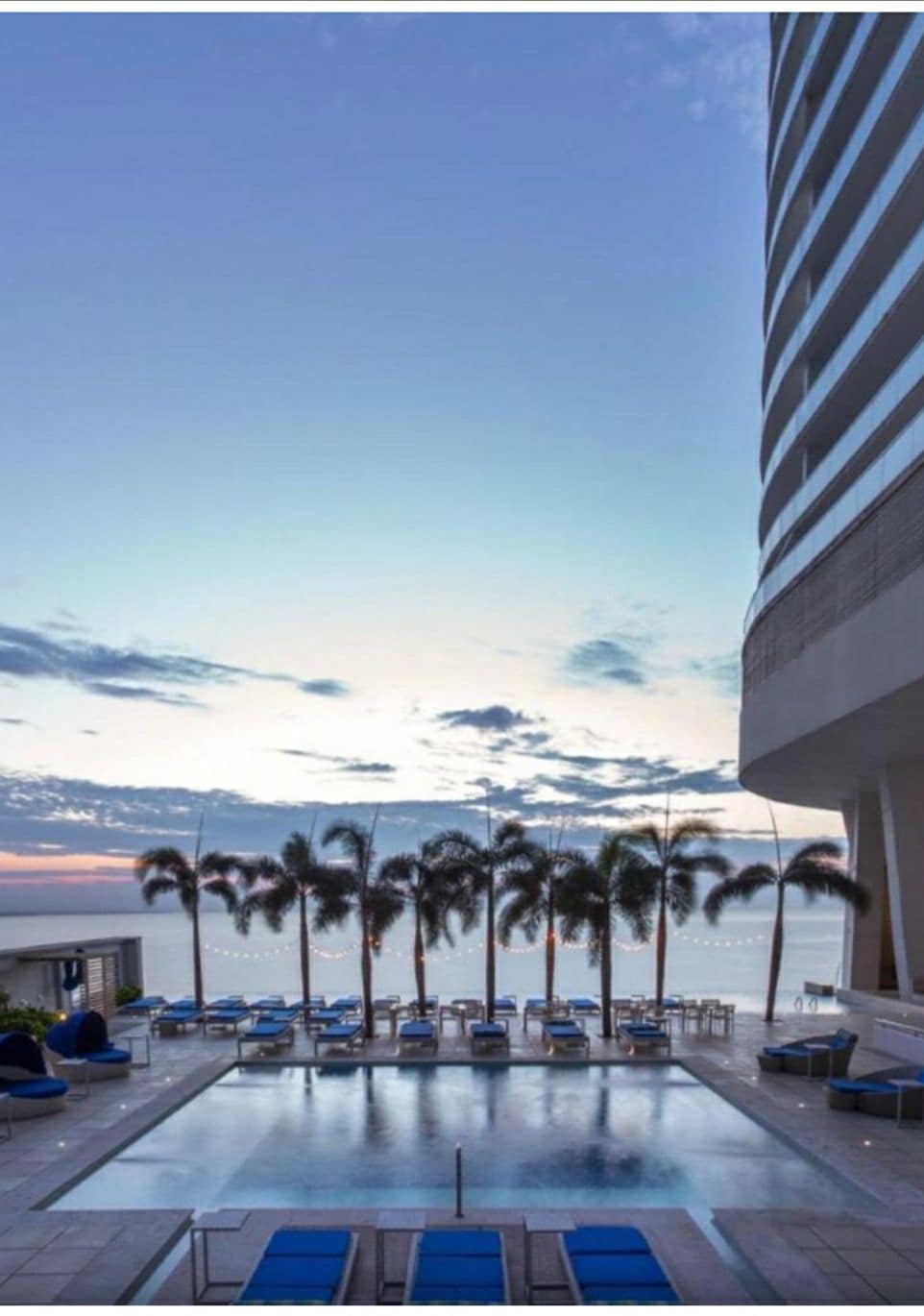 An outdoor pool area is surrounded by lounge chairs, with palm trees lining the perimeter. The twilight sky presents soft hues as the sun begins setting over the horizon, reflecting on the calm water surface.