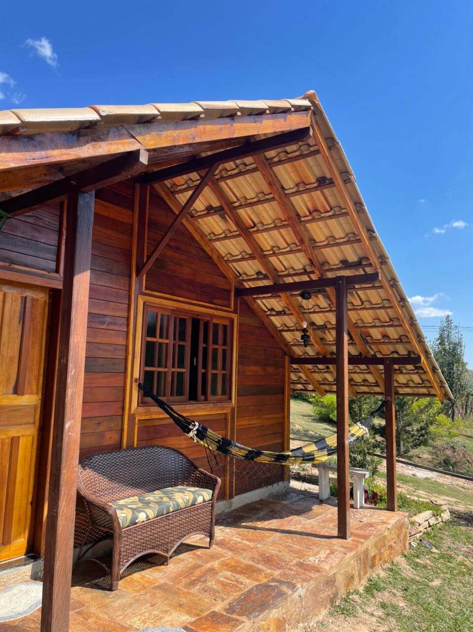 The exterior of a wooden cabin is shown, featuring a terracotta-tiled roof and large windows. A cozy sitting area includes a wicker chair and a hammock, situated on a stone patio, surrounded by greenery and blue sky.