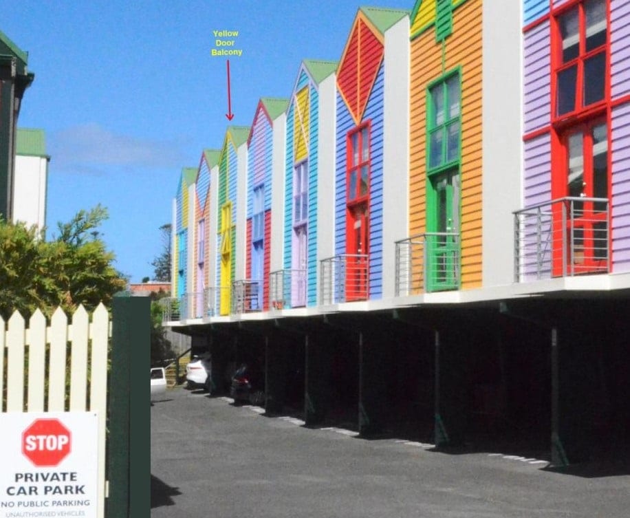 A row of colorful buildings is displayed, featuring vibrant facades in shades of green, yellow, and red. Each unit is supported by a sheltered parking area below, with a visible sign indicating private parking restrictions. A balcony with a yellow door is shown on one unit.
