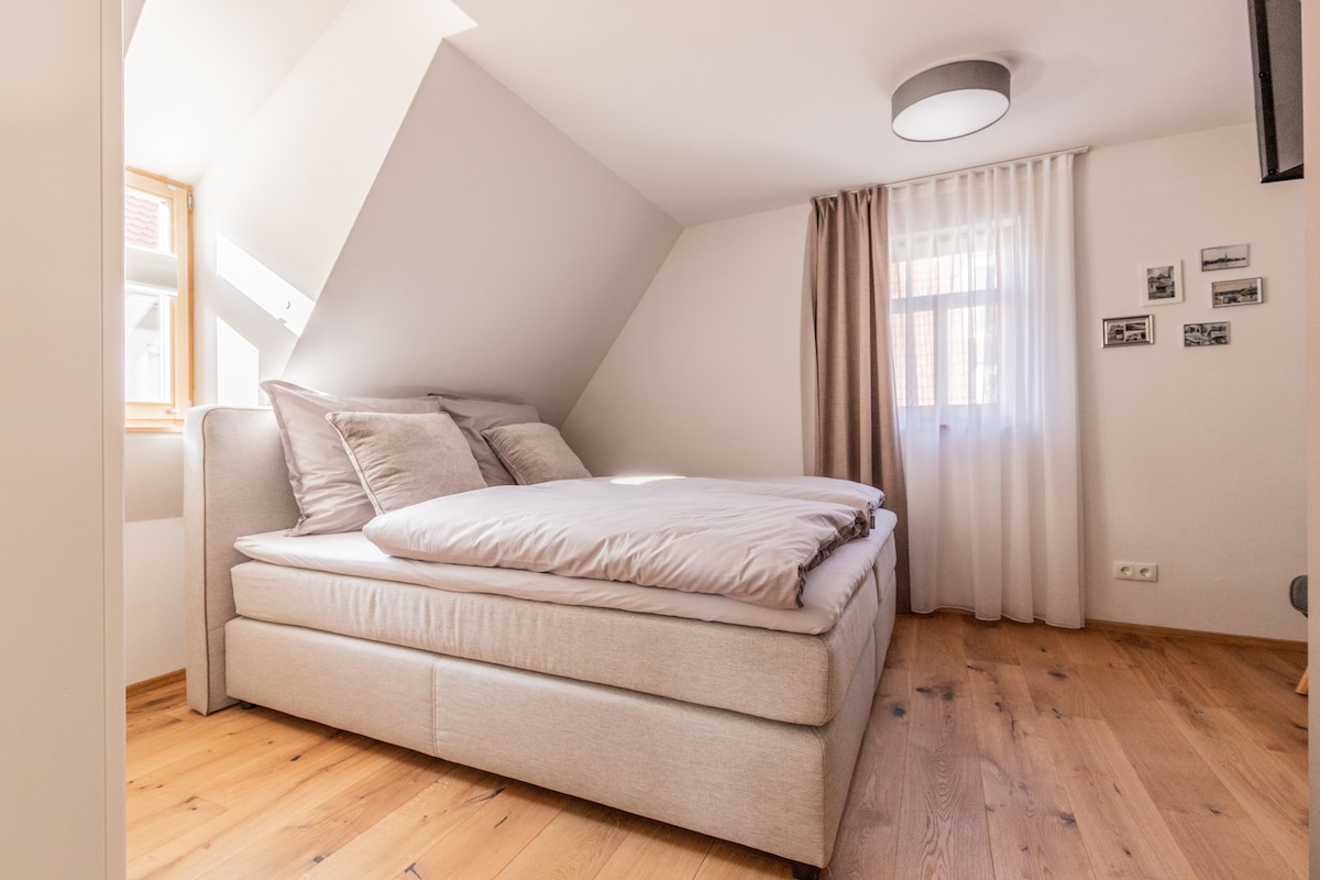 A comfortable sleeping area features a spacious bed dressed in soft linens. Natural light filters in through a window, complemented by sheer curtains. The room showcases light wooden flooring and light-colored walls, creating a calm and serene atmosphere.