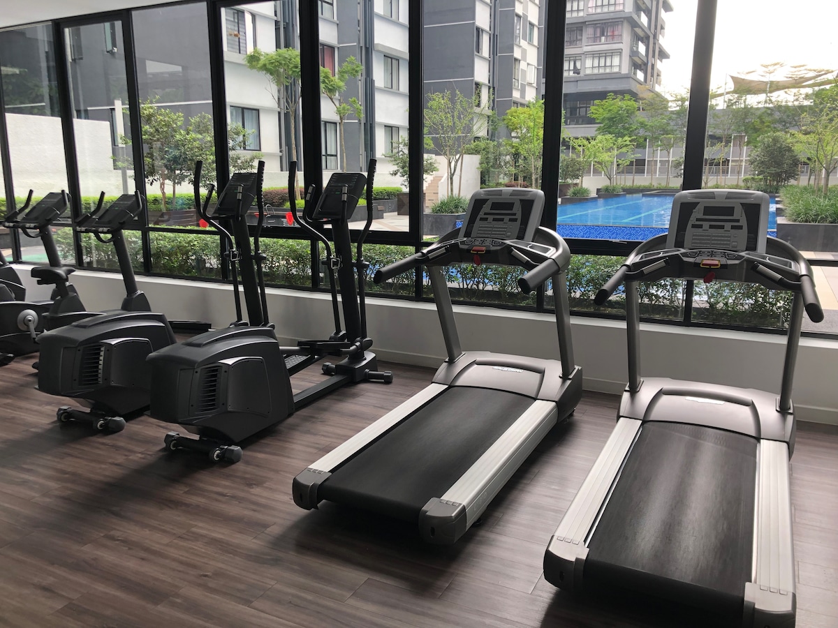 A fitness center features various exercise equipment, including treadmills and stationary bikes. Large windows allow natural light to illuminate the space, and views of greenery and a swimming pool are visible outside.
