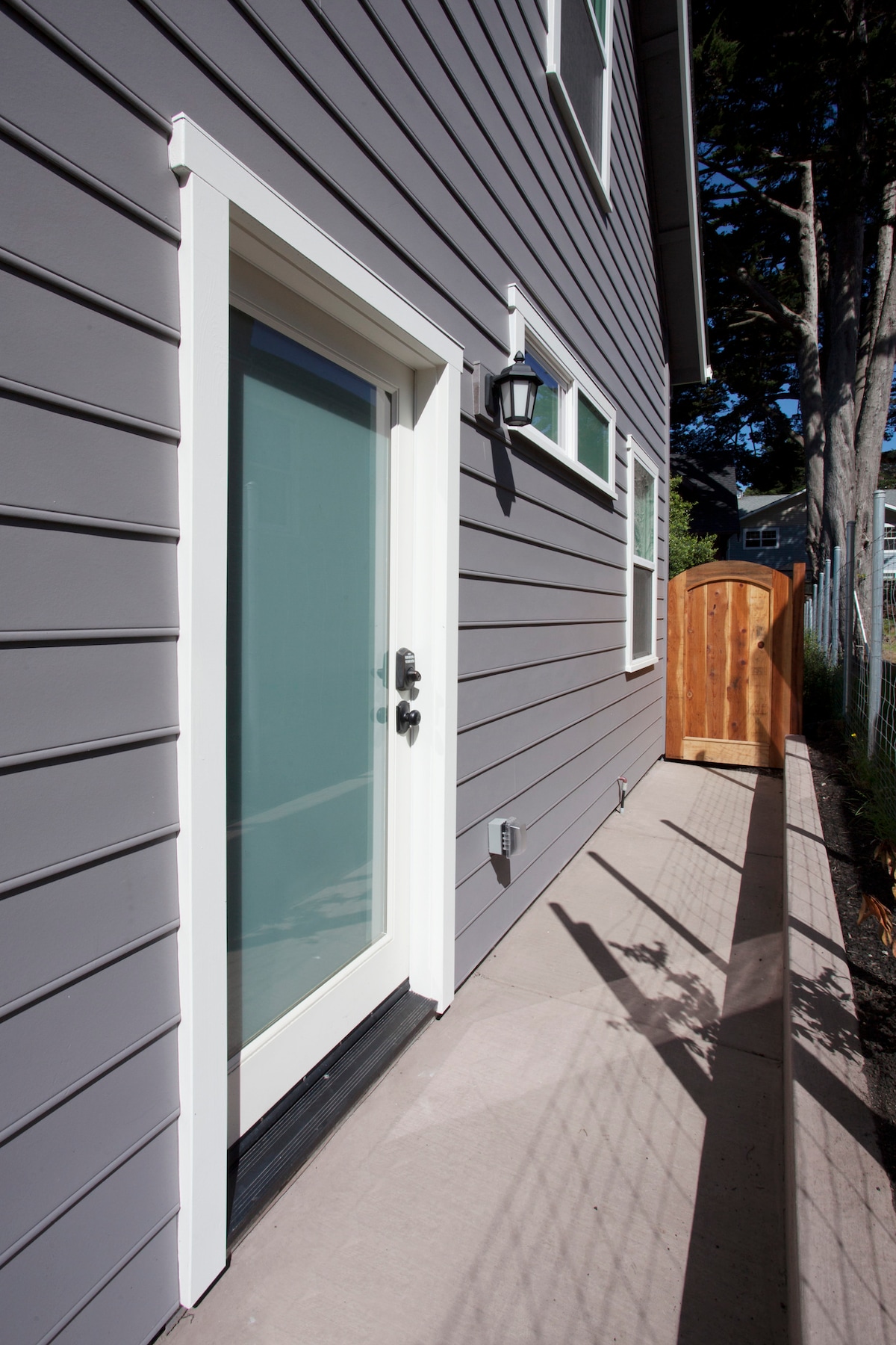 The image displays a private entrance to the studio, featuring a light-colored door with a modern handle. The exterior is clad in gray siding, complemented by white trim. A narrow walkway is visible beside the building, leading to a wooden gate and framed by a fence.