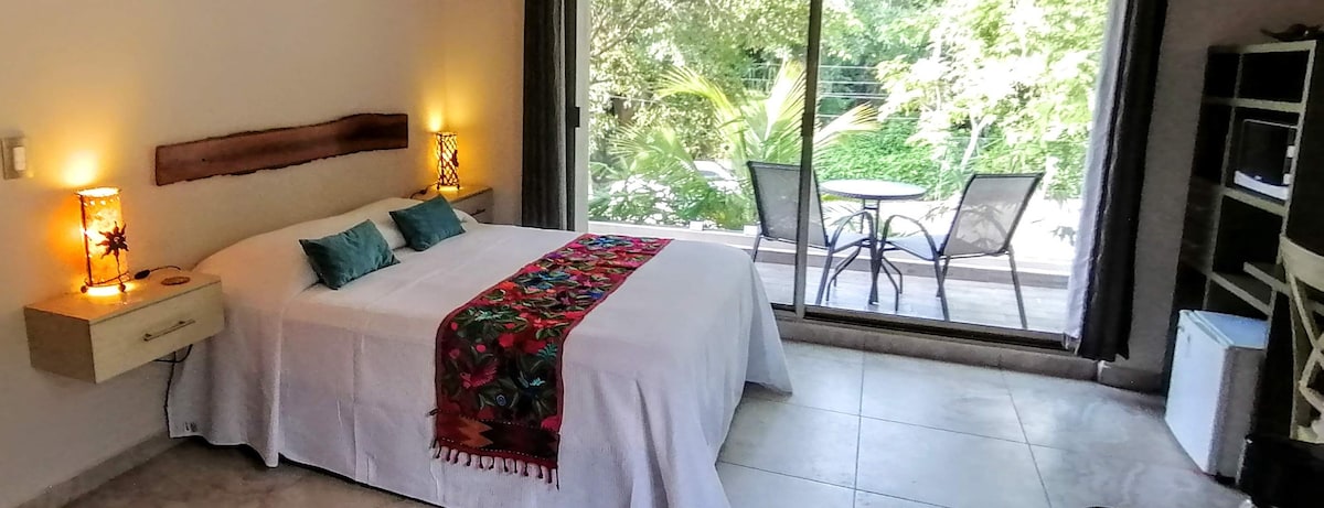 A spacious sleeping area features a large bed adorned with a colorful throw. Two bedside lamps illuminate the room, complemented by the natural light streaming through large glass doors that open to a private patio with a table and chairs overlooking greenery.