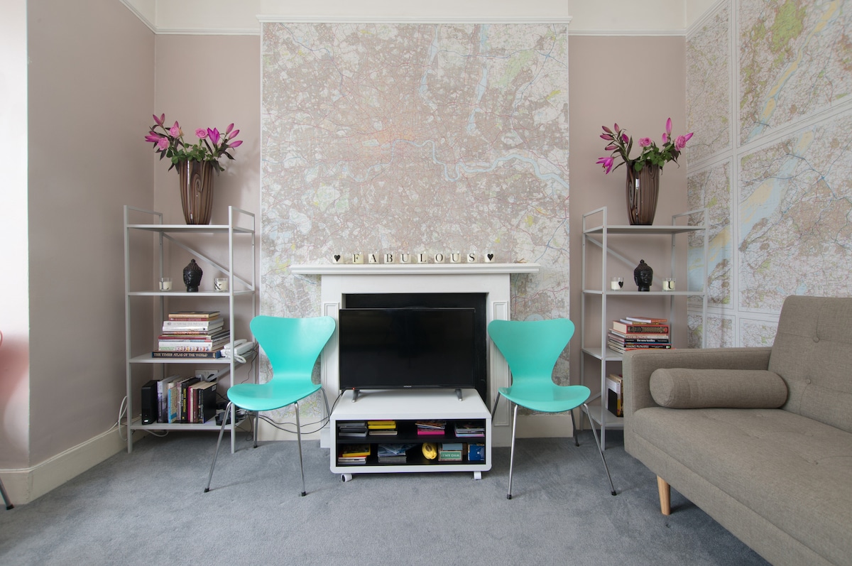 The living room features a decorative wall adorned with maps, complemented by two aqua armchairs and a neutral-toned sofa. A television is placed on a media console beneath the mantelpiece, which holds decorative items and bookshelves on either side filled with various books.