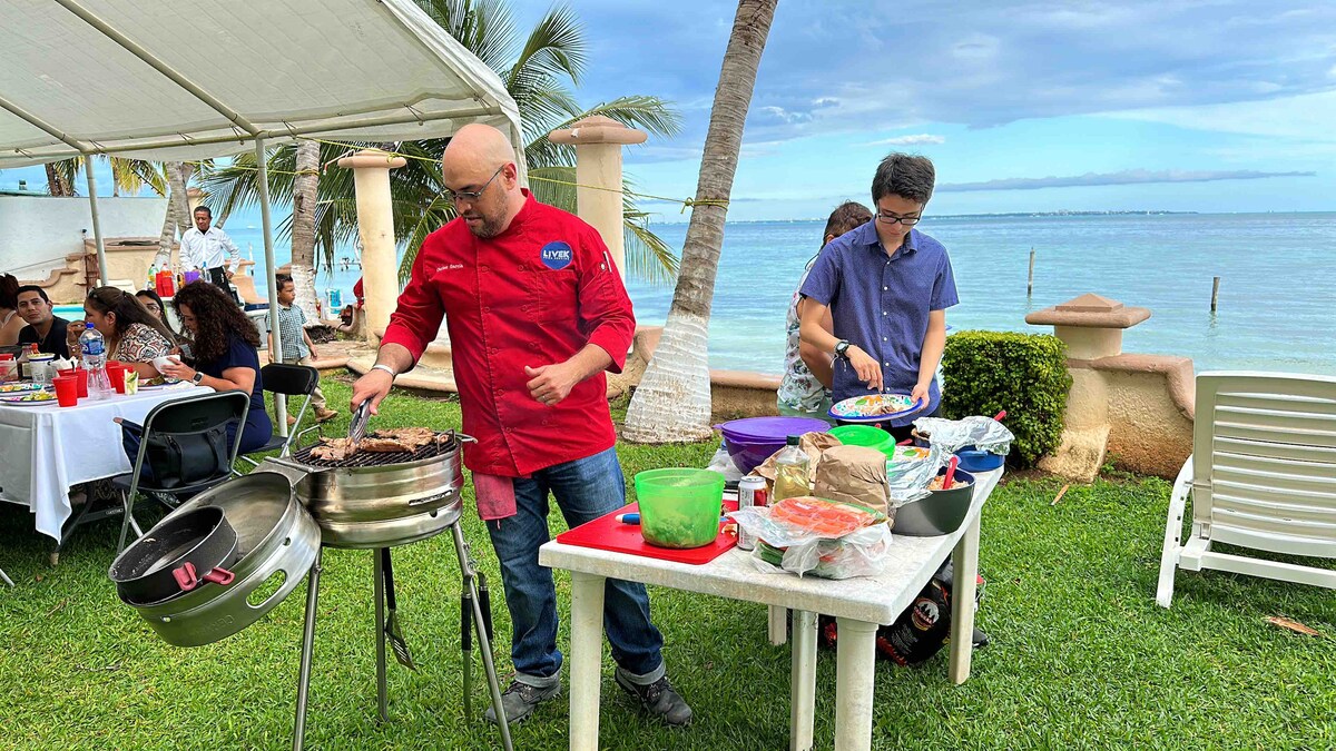 An outdoor cooking area is set near the water, featuring a grill with marinated food being prepared. A table displays colorful ingredients and utensils. Surrounding palm trees and neatly trimmed grass create a relaxed atmosphere, with dining tables visible in the background.