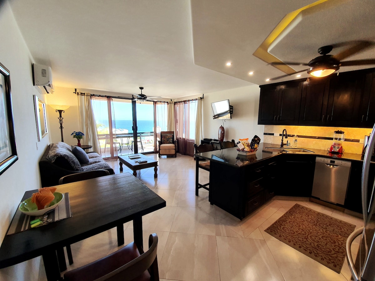 A bright living space features an open layout with a comfortable seating area and a dining table. Large glass doors lead to a balcony showcasing sweeping ocean views. The kitchen, equipped with modern appliances, complements the inviting ambiance of the suite.