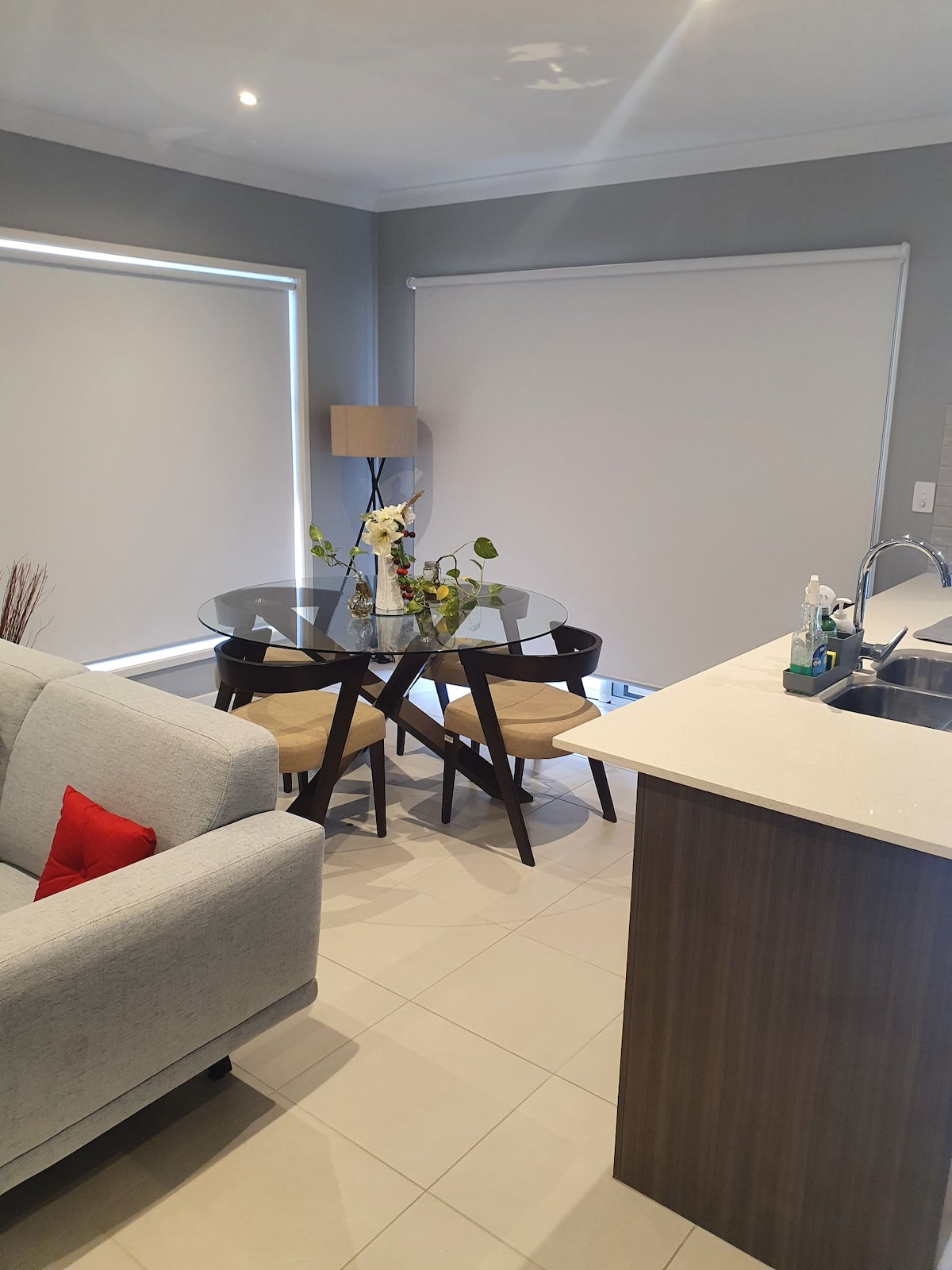 A modern living area showcases a glass dining table surrounded by two wooden chairs, accented with a vase of fresh flowers. A light-colored sofa is positioned nearby, while roller blinds cover the windows, allowing for light control.