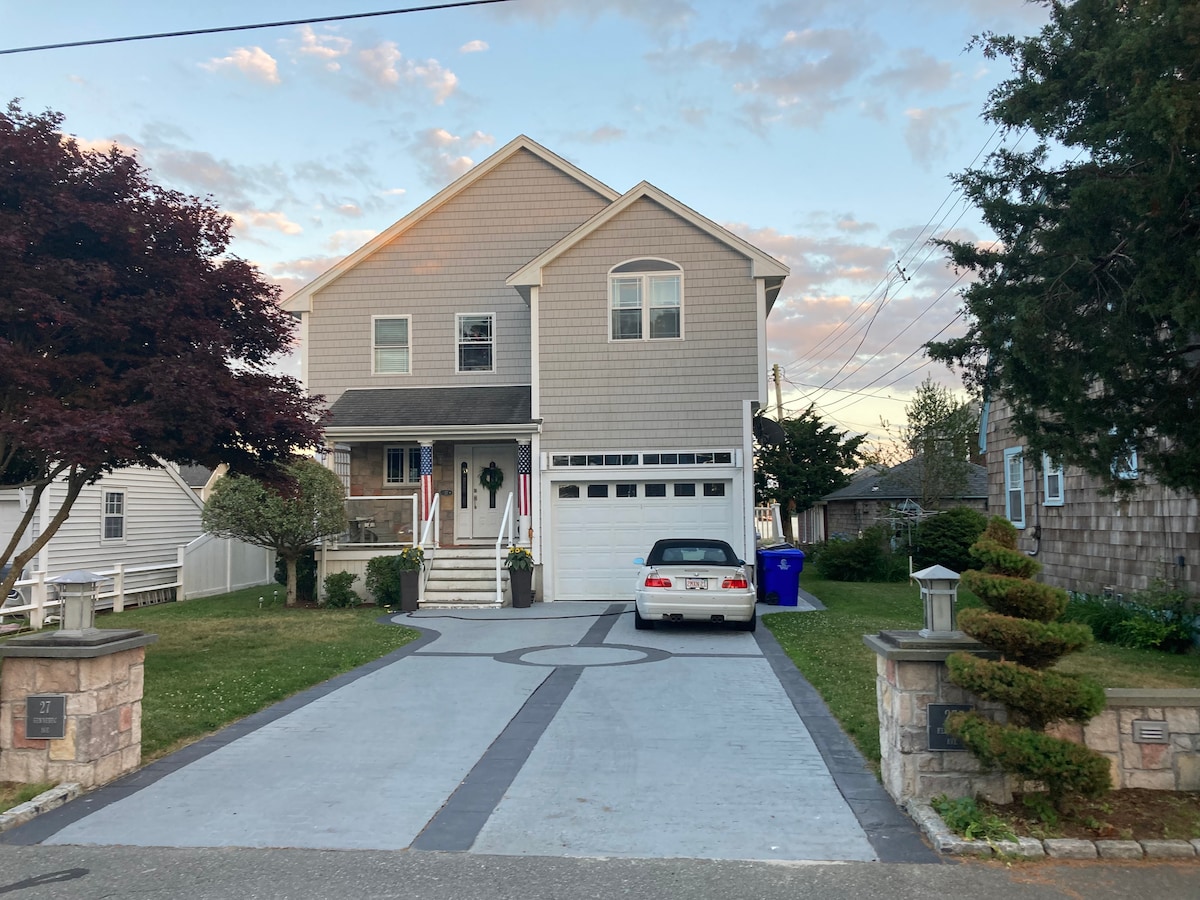 A modern two-story home is depicted, featuring a well-maintained front yard and a paved driveway. A light blue car is parked in front of the house, which is flanked by manicured shrubbery. The welcoming entryway is complete with steps leading to the porch.