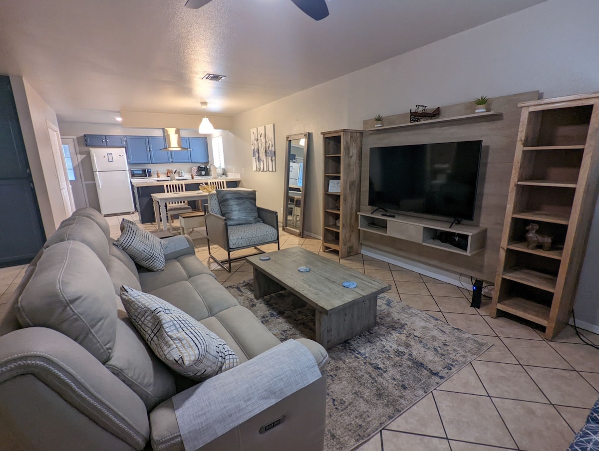 A spacious living area is visible, featuring a light-colored sectional sofa, a wooden coffee table, and a large television mounted on a media console. Neutral tones are complemented by a patterned area rug, with dining seating and a kitchen visible in the background.
