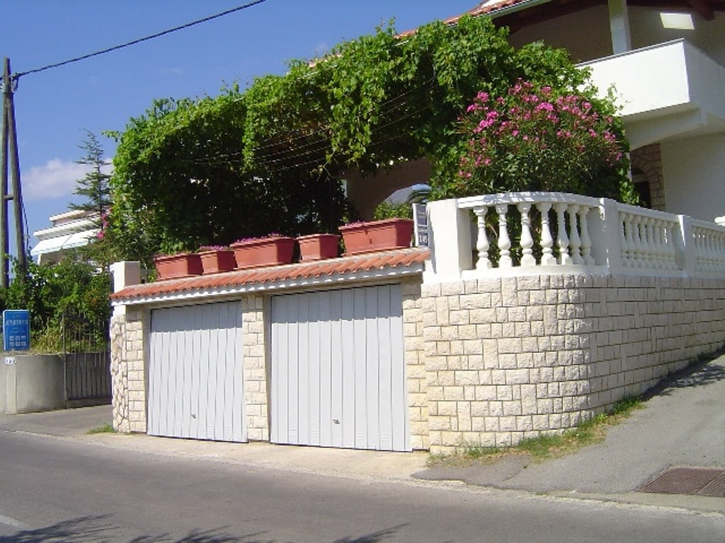 A two-garage structure is visible at street level, surrounded by a stone wall. The upper level features a balcony adorned with green foliage and flowerpots, providing a vibrant touch to the exterior. The sunny sky adds a bright backdrop to the scene.