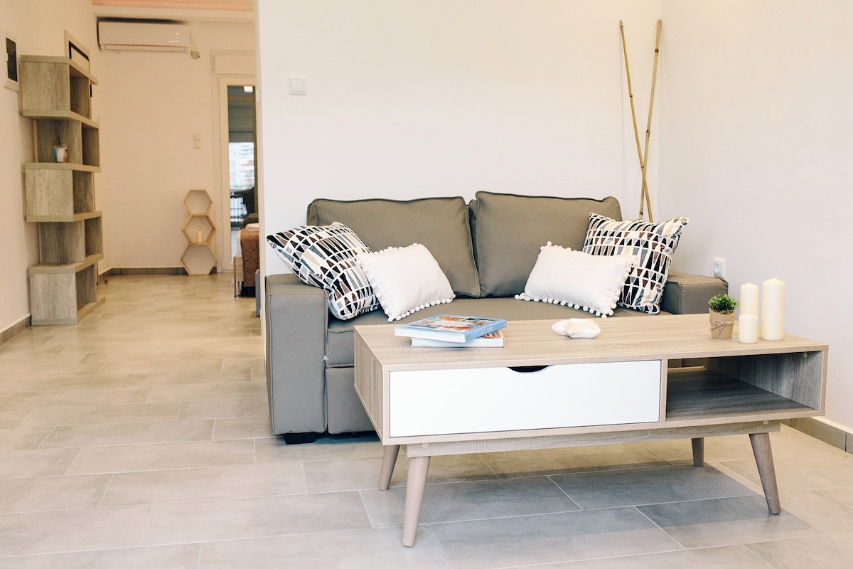 A light-filled living area features a gray couch adorned with black and white patterned pillows. A modern coffee table with a drawer holds a magazine and a decorative item. Wooden shelves are visible, providing additional storage and a minimalist aesthetic.
