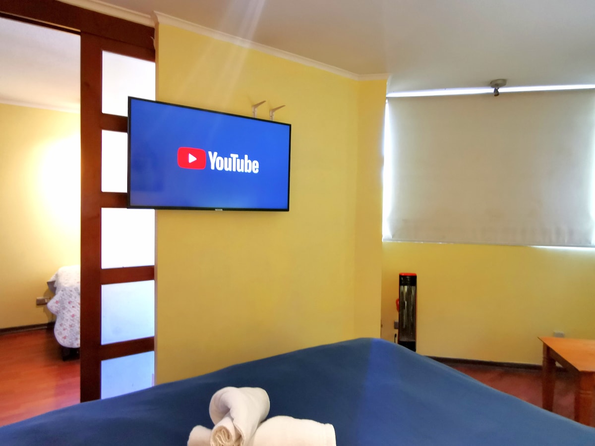 A modern bedroom features a spacious bed covered in a blue bedspread. A flat-screen television mounted on the wall displays the YouTube logo. Natural light enters through a nearby window partially covered by a light shade. A small table is visible in the corner.