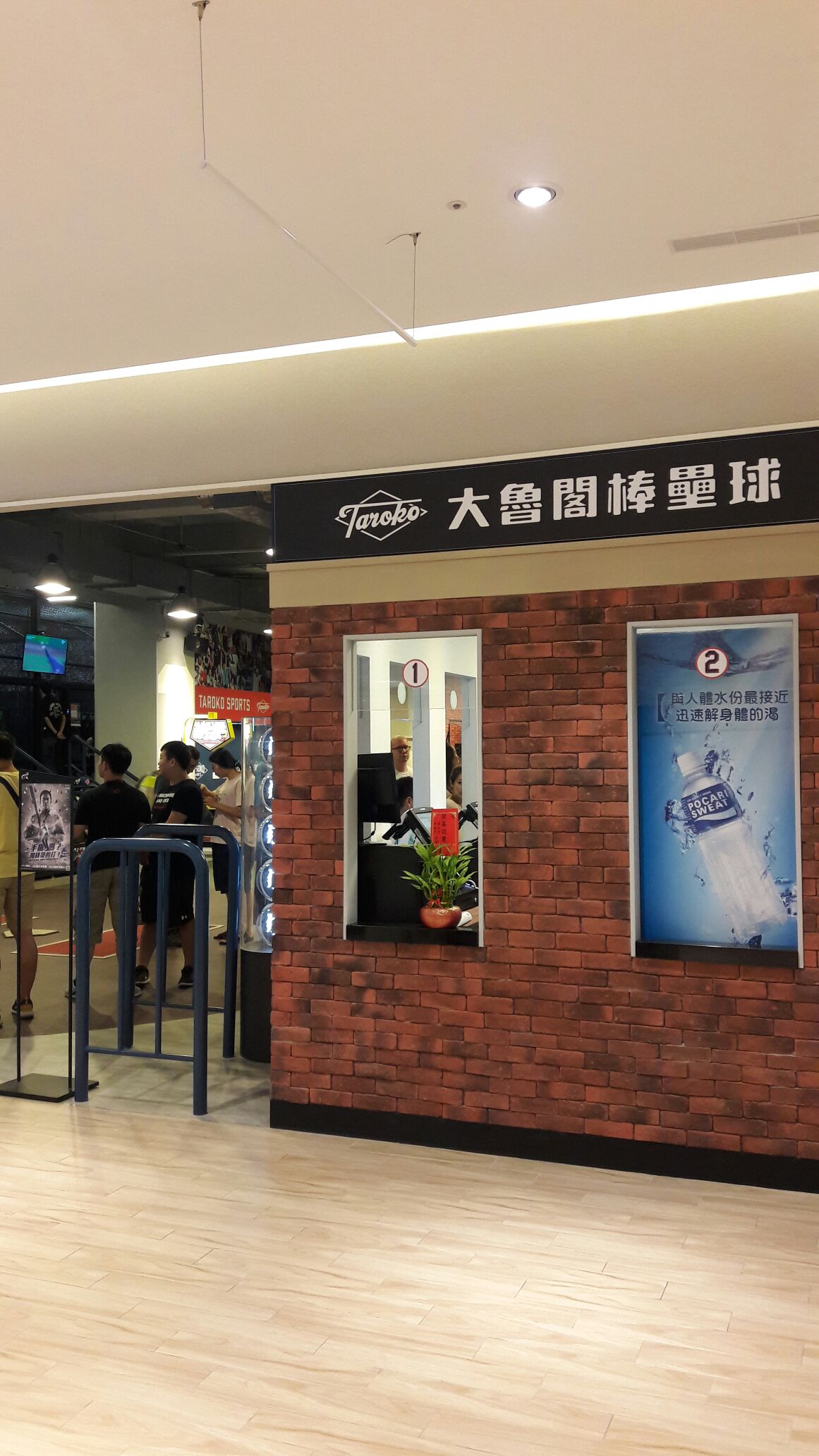 An entrance to a recreational area features a brick wall design with a sign displaying the name in large characters. A spacious area can be seen inside, with people engaging in various activities. There are potted plants near the entrance, adding a touch of greenery.